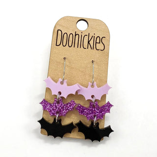Three pairs of bat-shaped earrings in pink, purple glitter, and black on a display card.